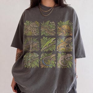 Op de afbeelding: Een antracietgrijs T-shirt met korte mouwen voorzien van een raster van negen gedetailleerde illustraties van diverse reptielen en amfibieën, waaronder slangen, schildpadden, een groene hagedis, een baardagame en een kameleon.