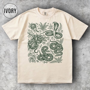 Puede incluir: Camiseta de color marfil con una ilustración detallada de varias serpientes y follaje en tonos verdes. El diseño incluye una cobra, serpientes enroscadas y elementos botánicos. La palabra "IVORY" está en un círculo negro en la parte superior izquierda.