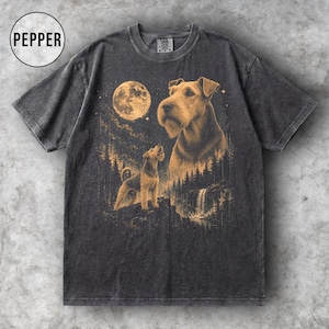 Peut inclure: Un t-shirt gris anthracite à manches courtes avec une impression graphique représentant deux terriers Airedale dans un paysage forestier sous une pleine lune. Le motif inclut une cascade et un ciel étoilé. Le mot PEPPER est inscrit dans un cercle.