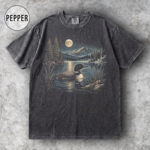 May include: A charcoal grey short-sleeve t-shirt featuring a graphic print of a loon swimming on a lake at night. The scene includes a full moon, pine trees, and distant mountains. The word PEPPER is printed in a circle in the top left corner.