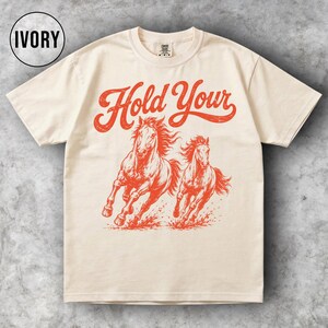 May include: Ivory-colored t-shirt featuring a red graphic of two running horses with the text "Hold Your" in a vintage font. The shirt has a classic crew neck and short sleeves, suitable for casual wear.