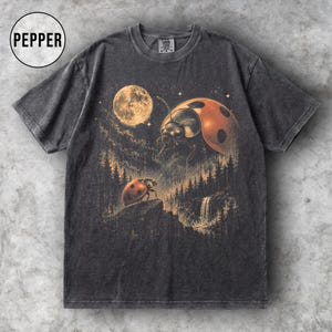 May include: Dark grey t-shirt with a surreal illustration of two ladybirds, a full moon, and a forest scene. The ladybirds are red and black. The word "PEPPER" is in a white circle in the upper left.