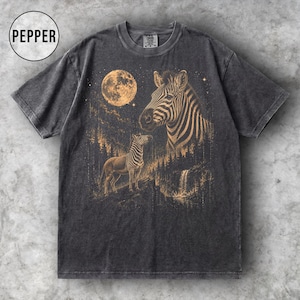 Può includere: Una t-shirt grigio antracite a maniche corte con una stampa grafica di una zebra in una foresta, una grande luna e una seconda testa di zebra più grande sullo sfondo. Il testo PEPPER è stampato in un cerchio nell'angolo in alto a sinistra.