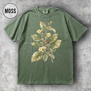 K&ouml;nnte beinhalten: Ein moosgr&uuml;nes T-Shirt mit einer botanischen Illustration eines bl&uuml;henden Zweigs, Bl&auml;ttern und einem Marienk&auml;fer. Das Wort "MOSS" befindet sich in einem wei&szlig;en Kreis. Das T-Shirt hat einen Rundhalsausschnitt und kurze &Auml;rmel.