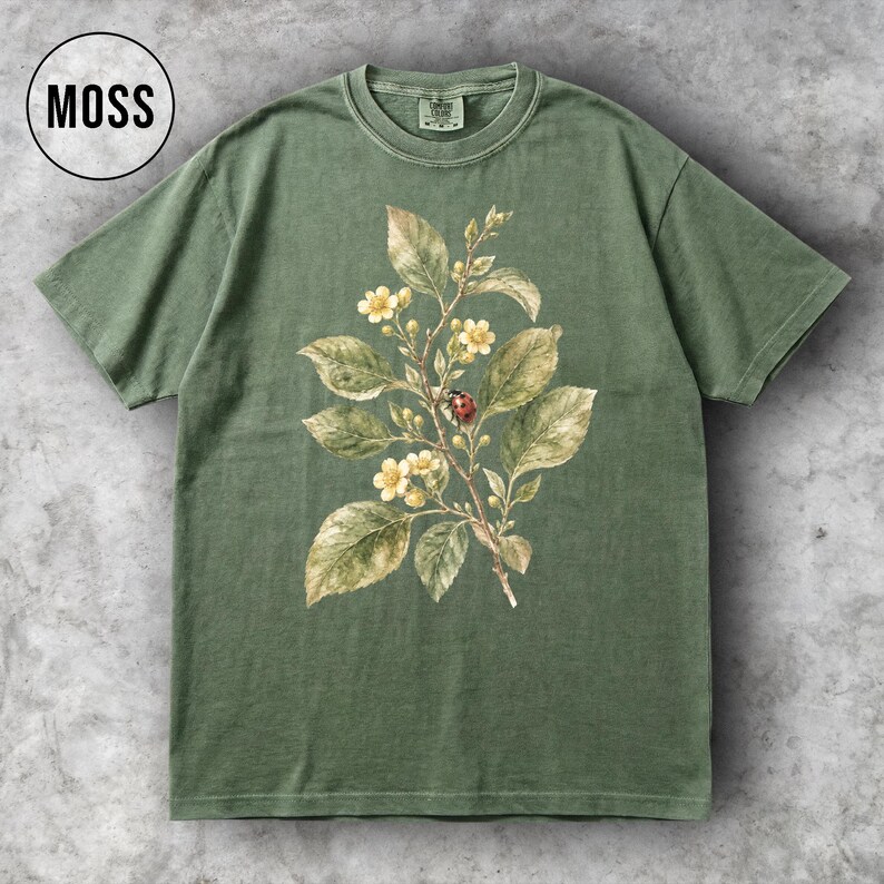 K&ouml;nnte beinhalten: Ein moosgr&uuml;nes T-Shirt mit einer botanischen Illustration eines bl&uuml;henden Zweigs, Bl&auml;ttern und einem Marienk&auml;fer. Das Wort "MOSS" befindet sich in einem wei&szlig;en Kreis. Das T-Shirt hat einen Rundhalsausschnitt und kurze &Auml;rmel.