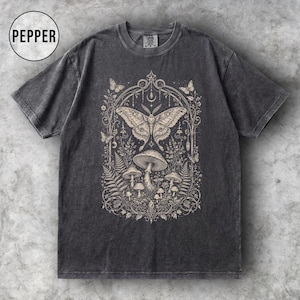 May include: Dark gray t-shirt featuring a detailed illustration of a moth, mushrooms, and foliage within an arched frame. The design is in a neutral color palette, creating a vintage aesthetic. The word "PEPPER" is in a circle in the upper left corner.