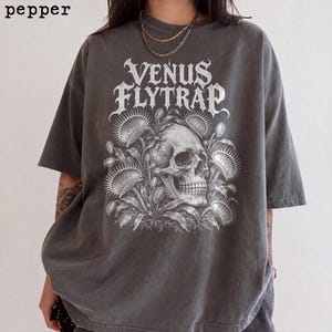Può includere: Una t-shirt a maniche corte color grigio antracite con una stampa grafica bianca raffigurante un teschio umano circondato da piante carnivore dionee. Il testo 'VENUS FLYTRAP' è stampato con un carattere stilizzato sopra il teschio.