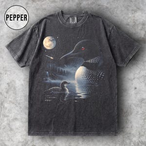 May include: Dark grey t-shirt with a detailed illustration of a loon on a lake under a starry night sky with a full moon. The design includes a smaller loon in the water and a waterfall in the background. The word "PEPPER" is in a circle in the upper left.