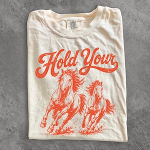 May include: Cream-colored t-shirt with the words "Hold Your" in orange script above a graphic of two running horses. The shirt is folded and has a Comfort Colors tag.