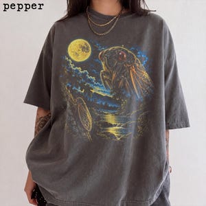 以下が含まれることがあります： チャコールグレーのTシャツで、満月の下の川の近くに止まる大きなセミのグラフィックプリントが施されています。デザインには木の枝に止まる小さなセミも含まれており、青と黄色のハイライトが特徴です。黒字でpepperという文字がプリントされています。