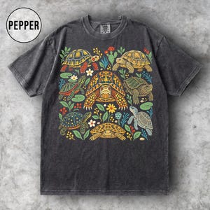 Könnte beinhalten: Ein anthrazitfarbenes Kurzarm-T-Shirt mit einem Grafikdruck verschiedener Schildkröten, umgeben von bunten Blumen und grünen Blättern. Der Text PEPPER ist in einem schwarzen Kreis in der oberen linken Ecke des Bildes aufgedruckt.