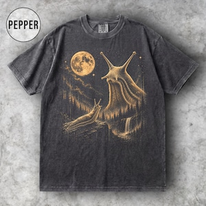 May include: A charcoal grey short-sleeve t-shirt featuring a graphic print of two large snails in a forest landscape under a full moon. The design includes a waterfall and starry sky. The word PEPPER is printed in a circle in the top left corner.