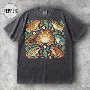 Puede incluir: Una camiseta gris carbón de manga corta con un estampado gráfico central de seis sapos de varios colores en tonos naranja, verde y marrón, rodeados de pequeñas hojas, flores y bellotas de colores. La palabra PEPPER está impresa en un círculo.