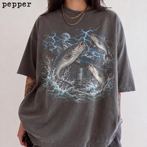 Puede incluir: Una camiseta gris carbón de manga corta con un estampado gráfico de tres siluros saltando fuera del agua sobre un fondo de montañas, una luna llena y rayos azules. La palabra pepper está impresa en negro en la esquina superior izquierda.
