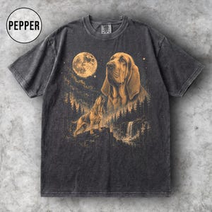 May include: A charcoal grey short-sleeve t-shirt featuring a graphic print of a bloodhound, a full moon, a forest, and a waterfall in shades of gold. The text PEPPER is printed in a circle in the upper left corner of the garment.