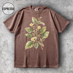 K&ouml;nnte beinhalten: Braunes T-Shirt mit botanischem Aufdruck. Das Design zeigt gr&uuml;ne Bl&auml;tter, gelbe Bl&uuml;ten und einen roten Marienk&auml;fer. Das T-Shirt hat einen Rundhalsausschnitt und kurze &Auml;rmel. Das Wort "ESPRESSO" befindet sich in einem Kreis.