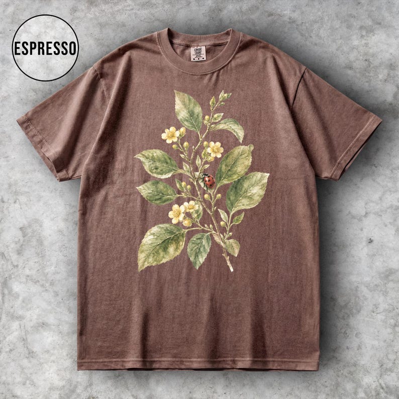 K&ouml;nnte beinhalten: Braunes T-Shirt mit botanischem Aufdruck. Das Design zeigt gr&uuml;ne Bl&auml;tter, gelbe Bl&uuml;ten und einen roten Marienk&auml;fer. Das T-Shirt hat einen Rundhalsausschnitt und kurze &Auml;rmel. Das Wort "ESPRESSO" befindet sich in einem Kreis.