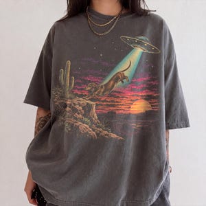 Może przedstawiać: Ciemnoszary t-shirt z krótkim rękawem z nadrukiem przedstawiającym jamnika unoszonego przez promień UFO. Scena zawiera pustynny krajobraz z kaktusem, zachodem słońca i fioletowym niebem. Koszulka ma okrągły dekolt i luźny krój.
