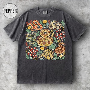 Könnte beinhalten: Ein anthrazitfarbenes Kurzarm-T-Shirt mit einem Grafikdruck verschiedener Schlangen in Rot-, Gelb-, Braun- und Schwarztönen, durchsetzt mit grünen Blättern und gelben Blumen. Der Text PEPPER ist in einem Kreis in der oberen linken Ecke gedruckt.