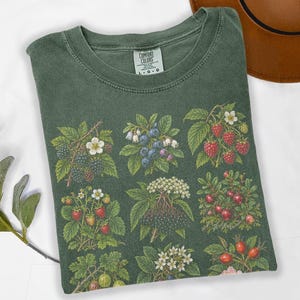 May include: A forest green crew neck t-shirt featuring a botanical print of various berries and flowers, including blackberries, blueberries, raspberries, strawberries, and elderflowers. The inner neck label displays the text Comfort Colors L G G.
