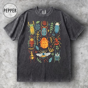 May include: Dark grey t-shirt with a colourful insect illustration. The design features various beetles and bugs in shades of yellow, orange, blue, and green, with floral accents. The word "PEPPER" is in a white circle in the upper left.