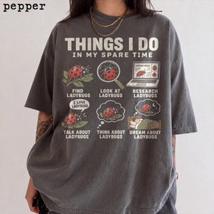 May include: A charcoal grey short-sleeve t-shirt featuring the text THINGS I DO IN MY SPARE TIME and six illustrations of ladybirds. The graphics include finding, looking at, researching, talking about, thinking about, and dreaming about ladybirds.