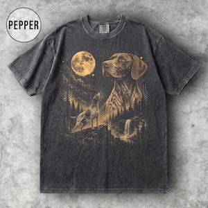 May include: A charcoal grey short-sleeve t-shirt featuring a graphic print of a dog looking at a full moon above a forest and waterfall. The design is rendered in shades of gold and tan. A circular badge in the top left corner contains the text PEPPER.