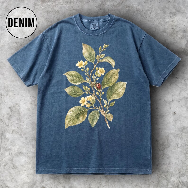 K&ouml;nnte beinhalten: Denimblaues T-Shirt mit einer botanischen Illustration. Das Design zeigt einen Zweig mit gr&uuml;nen Bl&auml;ttern, gelben Bl&uuml;ten und einem Marienk&auml;fer. Das Wort "DENIM" befindet sich in einem wei&szlig;en Kreis oben links.