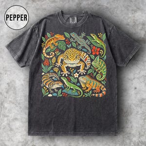 Könnte beinhalten: Ein anthrazitfarbenes Kurzarm-T-Shirt mit einem farbenfrohen Grafikdruck verschiedener Echsen, darunter ein Leopardgecko, ein Chamäleon und ein Leguan, umgeben von botanischen Elementen. Das Wort PEPPER ist in einem Kreis oben links aufgedruckt.