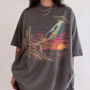 May include: A charcoal grey short-sleeve t-shirt featuring a graphic print of a German Shepherd dog being lifted by a UFO beam. The scene includes a desert landscape with a cactus, a rocky cliff, a sunset, and a starry purple and orange sky.
