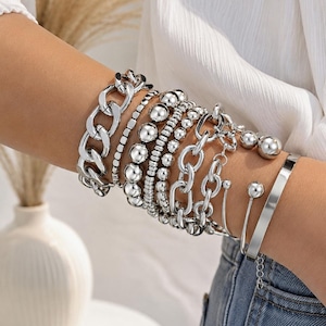 May include: A set of silver-toned bracelets featuring a variety of styles including a thick chunky chain, multiple strands of smooth round beads in varying sizes, a delicate oval link chain, and a sleek open cuff with spherical end caps. Virexia.