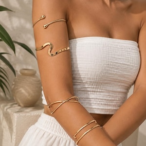 May include: A set of four gold-toned metal arm cuffs worn on the upper and lower arm. The set includes a simple open-ended cuff, a wavy design cuff, a crisscross cuff, and a double-banded cuff. Each piece features a smooth, polished finish.