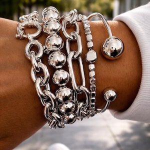 May include: An assortment of silver bracelets featuring different designs. The bracelets include chain links, beaded strands, and a cuff style with spherical details. The bracelets are worn on a wrist, displaying the various textures and styles.