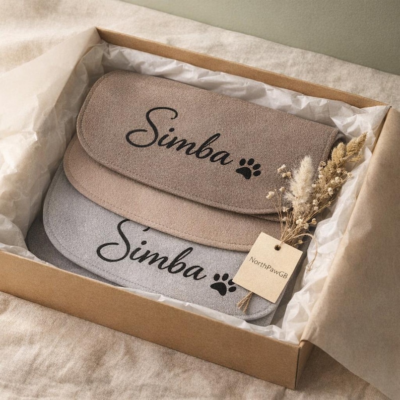 May include: A gift box containing three pet placemats in shades of beige and gray. Each placemat is oval-shaped and features the name "Simba" in cursive and a paw print. A small tag with text is attached to a bundle of dried flowers.