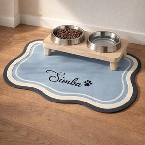 May include: A pet feeding station featuring two stainless steel bowls on a light wood stand. The stand rests on a blue mat with the name "Simba" and a paw print. The mat has a wavy border with blue, white, and dark grey accents.
