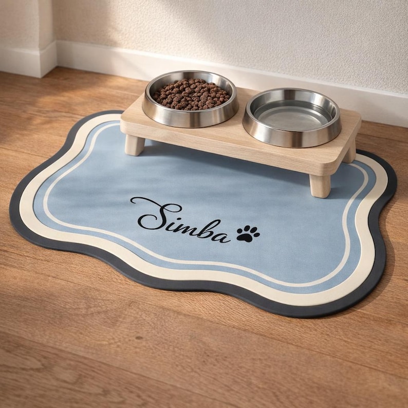 May include: A pet feeding station featuring two stainless steel bowls on a light wood stand. The stand rests on a blue mat with the name "Simba" and a paw print. The mat has a wavy border with blue, white, and dark grey accents.