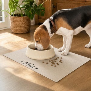 Peut inclure: Un tapis de nourriture pour animal de compagnie rectangulaire beige à surface texturée portant le nom Mia et de petites icônes de pattes en noir. Un bol rond côtelé de couleur crème est posé sur le tapis, rempli de croquettes brunes, avec un beagle.