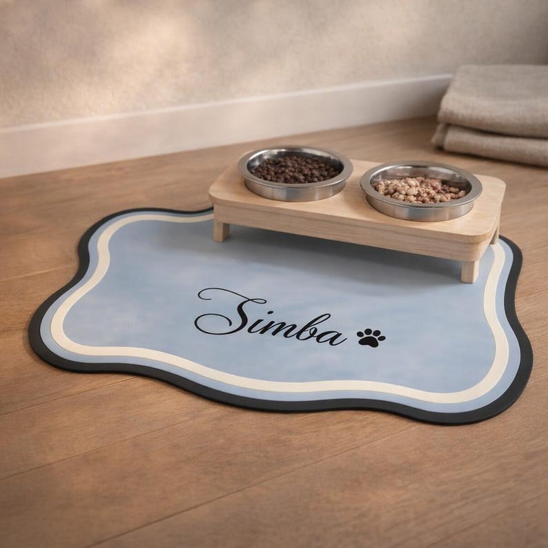 May include: A light blue pet food mat with a black border and the name "Simba" in script. The mat has a paw print and a wooden stand holding two stainless steel bowls filled with pet food.