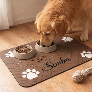 Könnte beinhalten: Ein Hund frisst aus einer beigen Schüssel auf einer braunen Matte mit weißen Pfotenabdrücken und dem Namen "Simba". Eine zweite Schüssel ist mit Hundefutter gefüllt. Ein beiges Seilspielzeug befindet sich in der unteren rechten Ecke.