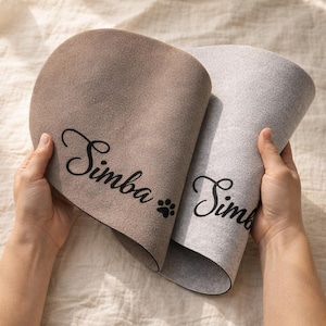 May include: Two pet placemats, one tan and one gray, are held up. The tan mat has the name "Simba" in black script with a paw print. The gray mat also has the name "Simba" in black script.