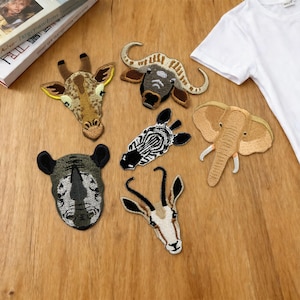 May include: Embroidered animal patches featuring a giraffe, buffalo, zebra, elephant, rhino, and gazelle. The patches are in various colors, including brown, black, white, and tan. The patches are arranged on a wooden surface.
