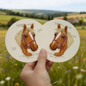 May include: Two circular coasters with embroidered horse head designs and red bridles. Text in English and Japanese includes "LEADSHOW" and "We love abundant nature." The background is a blurred countryside scene.
