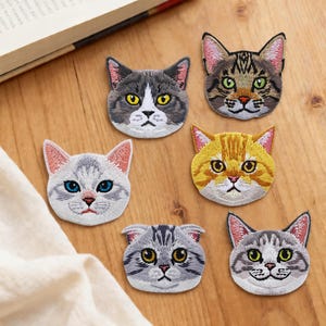 以下が含まれることがあります： さまざまな色と模様の、猫の顔が刺繍された6つのパッチのコレクション。 パッチは詳細なステッチが施されており、さまざまな品種とマーキングが施されています。 木の表面に配置され、背景には本と布があります。