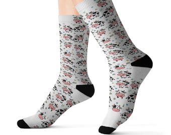 Könnte beinhalten: Ein Paar weiße Socken mit einem durchgehenden Muster aus Cartoon-Kühen mit schwarz-weißen Flecken und rosa Schnauzen. Die Socken haben einfarbige schwarze Fersen und Zehenbereiche, und die Kuh-Illustrationen sind mit kleinen blauen Sternen durchsetzt.
