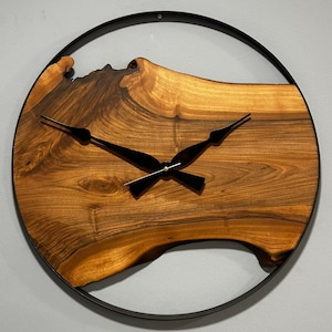Puede incluir: Un reloj de pared redondo con una esfera de madera natural y manecillas y marco de metal negro. La veta de la madera es visible y el reloj tiene un diseño moderno y minimalista. El diámetro del reloj es de aproximadamente 30 cm.