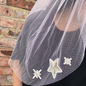 May include: A white sheer veil featuring a delicate Swiss dot pattern throughout the fabric. The bottom edge is decorated with three embroidered star-shaped appliqués, including one large central star covered in shimmering silver sequins.