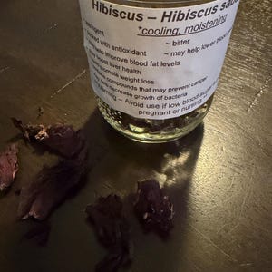 Puede incluir: Un tarro de cristal de té de hibisco con una etiqueta blanca. La etiqueta dice "Hibiscus - Hibiscus sabdariffa" y enumera propiedades como "refrescante, hidratante". Flores de hibisco secas están esparcidas alrededor del tarro sobre una superficie de madera oscura.