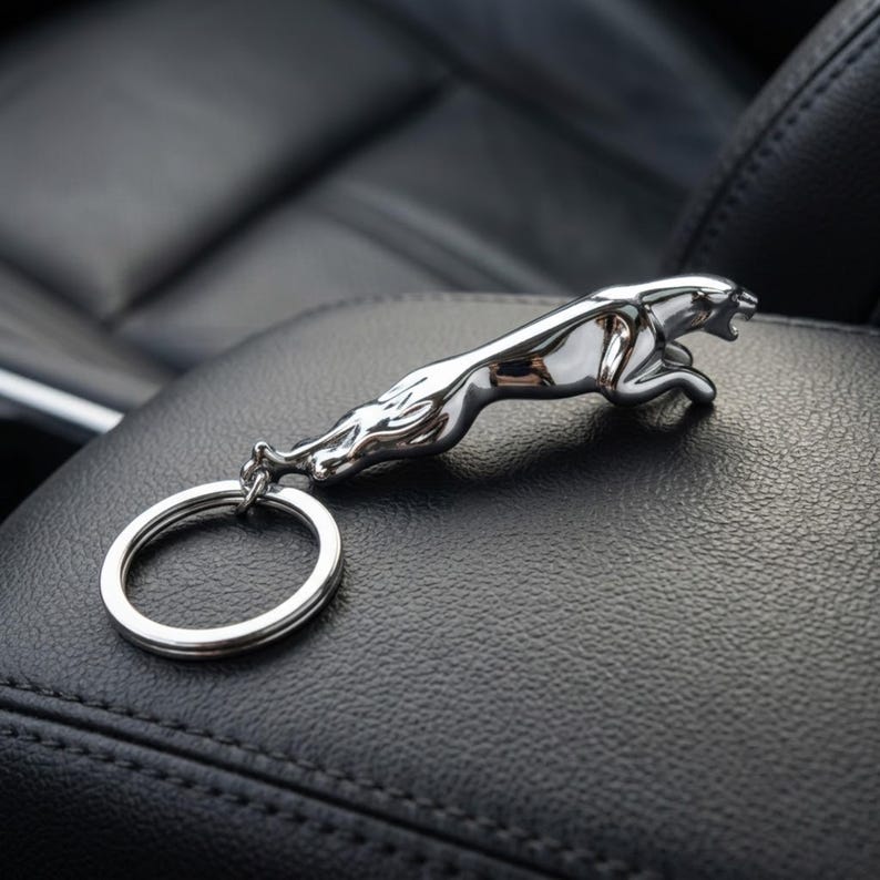May include: A chrome-finished Jaguar leaping cat keychain. The keychain features a detailed sculpture of the iconic car emblem, attached to a silver key ring. The keychain is resting on a black leather surface.