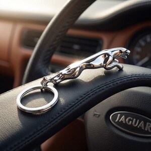 May include: A chrome Jaguar keychain featuring the iconic leaping jaguar emblem. The keychain is attached to a large silver ring and rests on a black leather steering wheel. The steering wheel has the Jaguar logo.