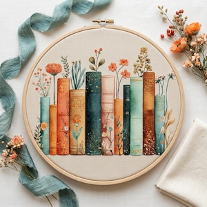 Puede incluir: Un bastidor de bordado de punto de cruz con un diseño de libros y flores. Los libros son de color naranja, verde azulado y dorado. Una cinta verde azulado y flores naranjas están dispuestas alrededor del bastidor.